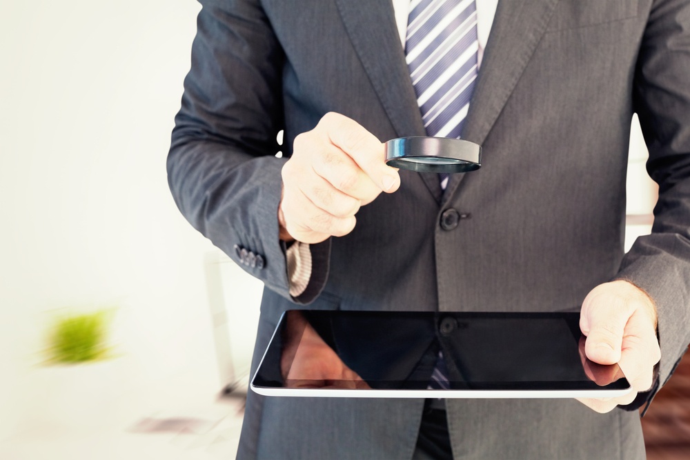 Businessman looking at tablet with magnifying glass against laptop on desk with glasses and notepad.jpeg